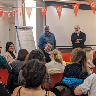 Two presenters standing at the front of a room, listening to feedback from their audience.