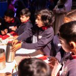 a young boy sits in a crowd of children, as he excitedly picks a story starter from a tin on the table in front of him.