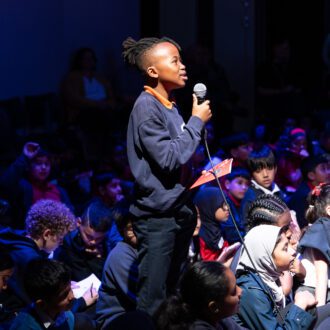 A young black boy stands proudly holding a microphone, addressing someone off camera. He is surrounded by other young children, sitting on the floor of a hall, who are looking up at him.