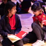 Two young girls sit on the floor writing in their notebooks, which are covered in stickers, chatting together.