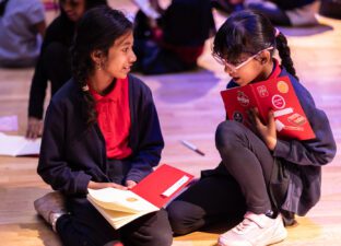 Two young girls sit on the floor writing in their notebooks, which are covered in stickers, chatting together.