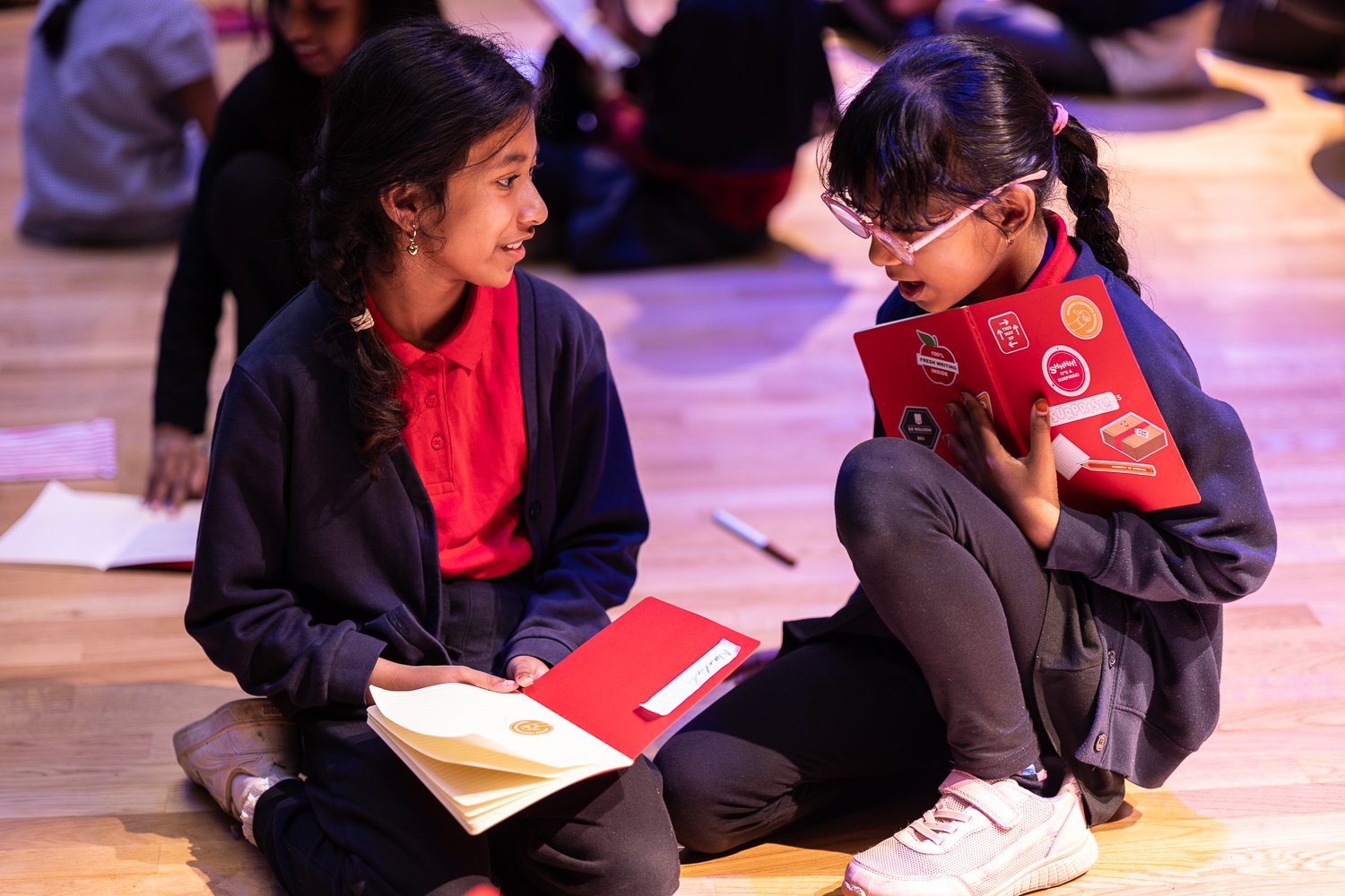 Two young girls sit on the floor writing in their notebooks, which are covered in stickers, chatting together.