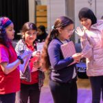 Four young girls smile and wave at the camera as they carry their notebooks and pens across the hall