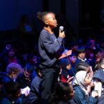 A young black boy stands proudly holding a microphone, addressing someone off camera. He is surrounded by other young children, sitting on the floor of a hall, who are looking up at him.