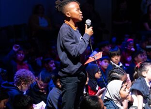 A young black boy stands proudly holding a microphone, addressing someone off camera. He is surrounded by other young children, sitting on the floor of a hall, who are looking up at him.