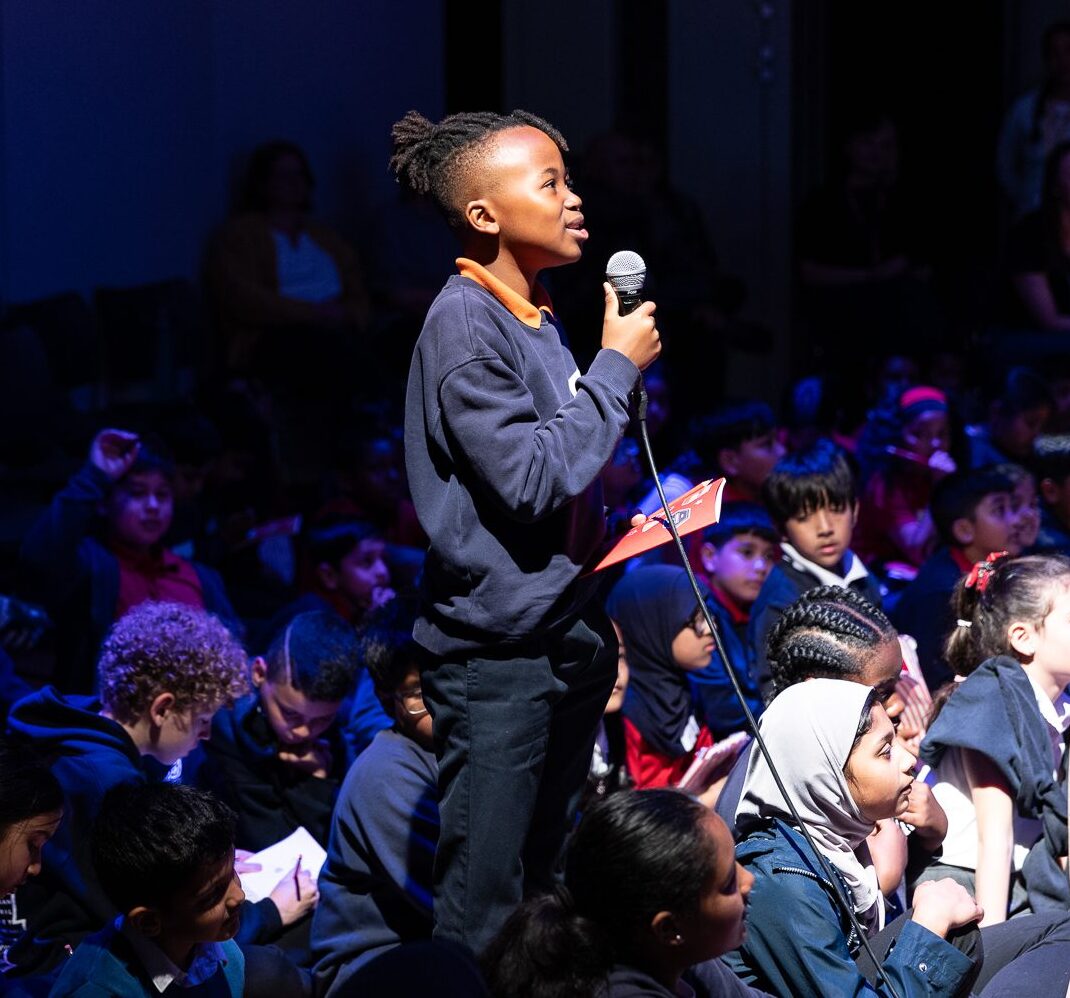 A young black boy stands proudly holding a microphone, addressing someone off camera. He is surrounded by other young children, sitting on the floor of a hall, who are looking up at him.