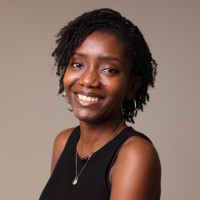 Keji, a black woman with short dark hair, sits smiling at the camera. She wears a black vest and gold necklace.