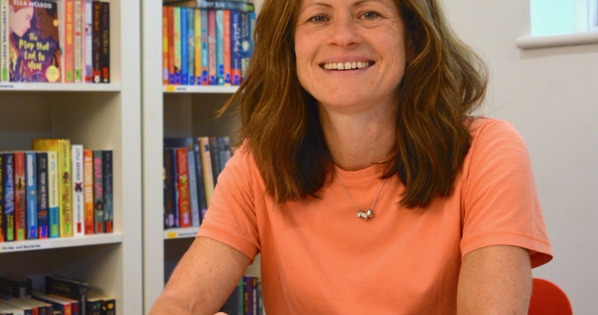 Laura - a white woman with long brown hair - sits smiling in front of a colourful bookshelf. Laura is wearing a peach t-shirt, a short silver necklace and a silver watch.