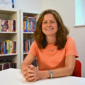 Laura - a white woman with long brown hair - sits smiling in front of a colourful bookshelf. Laura is wearing a peach t-shirt, a short silver necklace and a silver watch.