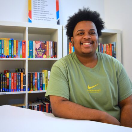 Ashante, a young black man, sits in a green t-shirt and dark hair smiling broadly at the camera.