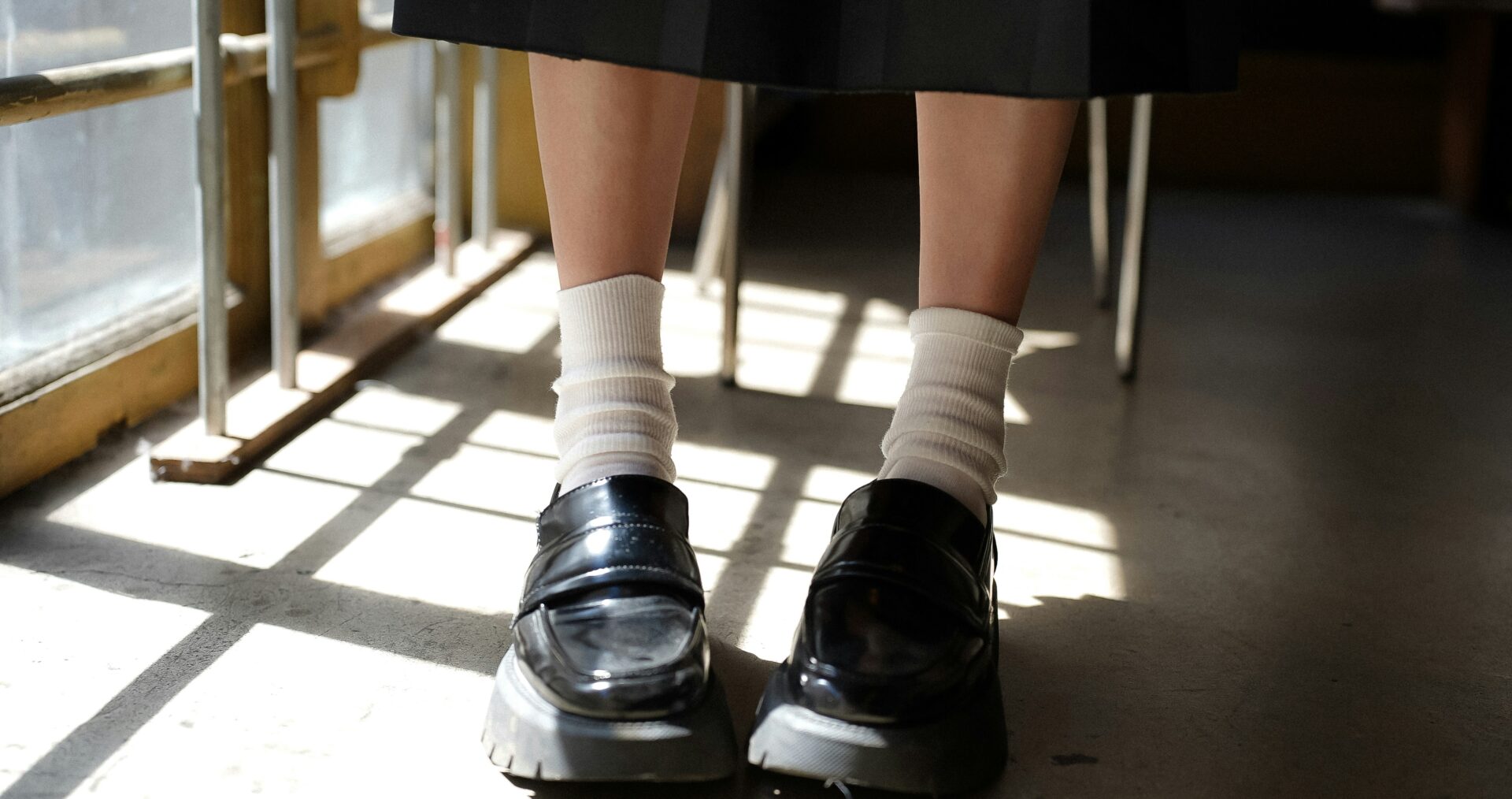 A close up image of a young person wearing short white socks and black school shoes, standing in a classroom with a desk behind them.