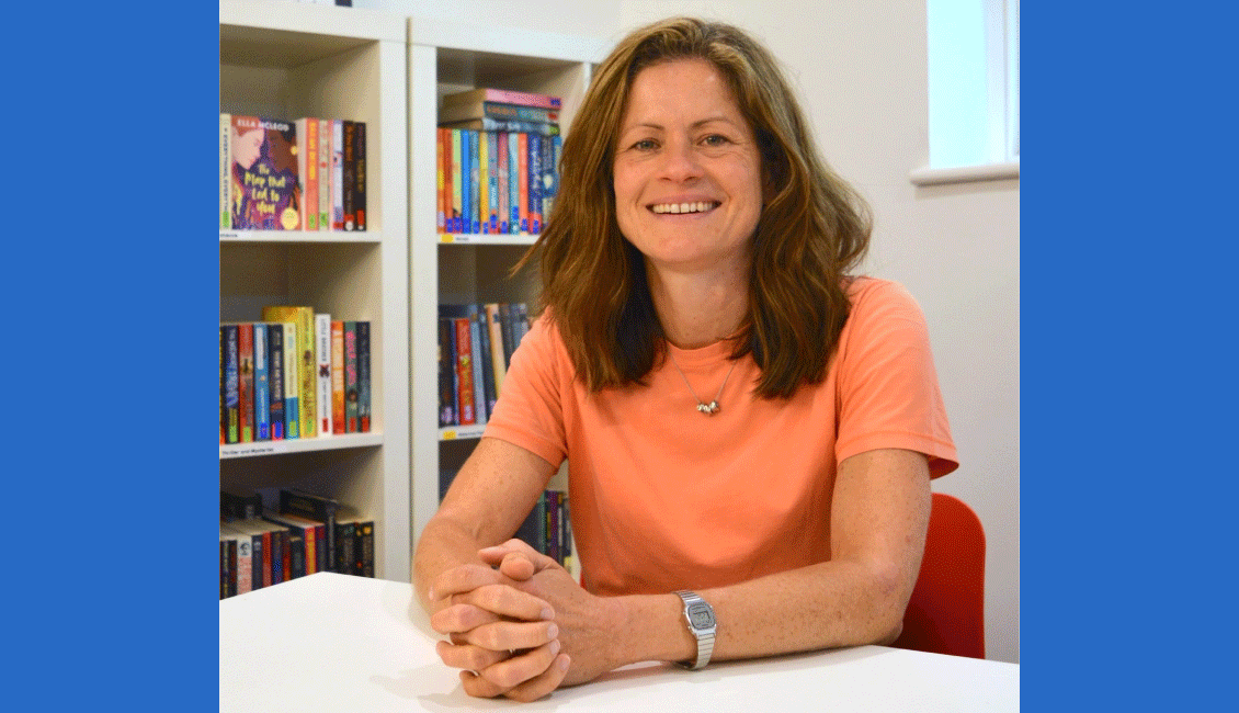 Laura - a white woman with long brown hair - sits smiling in front of a colourful bookshelf. Laura is wearing a peach t-shirt, a short silver necklace and a silver watch.