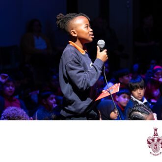 A young boy stands in the middle of a crowded room, speaking into a microphone.