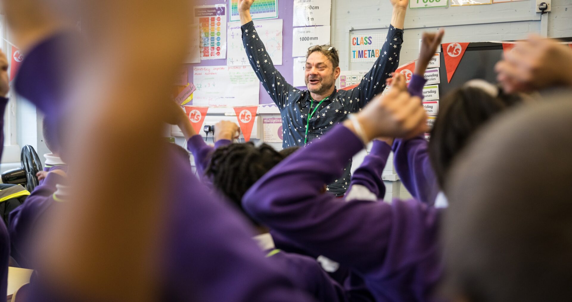 Justin - a writing facilitator with a purple shout, short hair and a big grin - lefts his arms up joyfully celebrating in front of a busy classroom with lots of happy and excited children, wearing purple school jumpers.