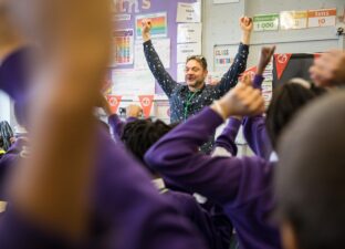 Justin - a writing facilitator with a purple shout, short hair and a big grin - lefts his arms up joyfully celebrating in front of a busy classroom with lots of happy and excited children, wearing purple school jumpers.