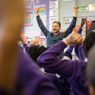 Justin - a writing facilitator with a purple shout, short hair and a big grin - lefts his arms up joyfully celebrating in front of a busy classroom with lots of happy and excited children, wearing purple school jumpers.