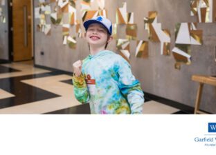 A young boy in colourful hat and hoodie pumps his fist in an excited, happy way.