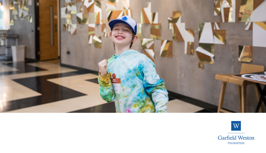 A young boy in colourful hat and hoodie pumps his fist in an excited, happy way.