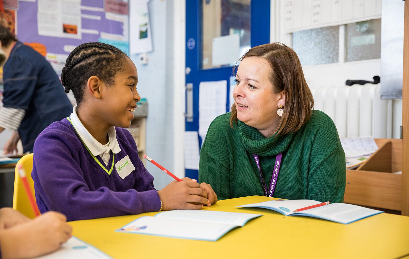 A female teacher in a green jumper sits with a student in a purple jumper as they write a story together