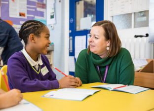 A female teacher in a green jumper sits with a student in a purple jumper as they write a story together