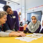 Three young people sit at a table with a writing facilitator smiling and laughing laying out words on strips of paper
