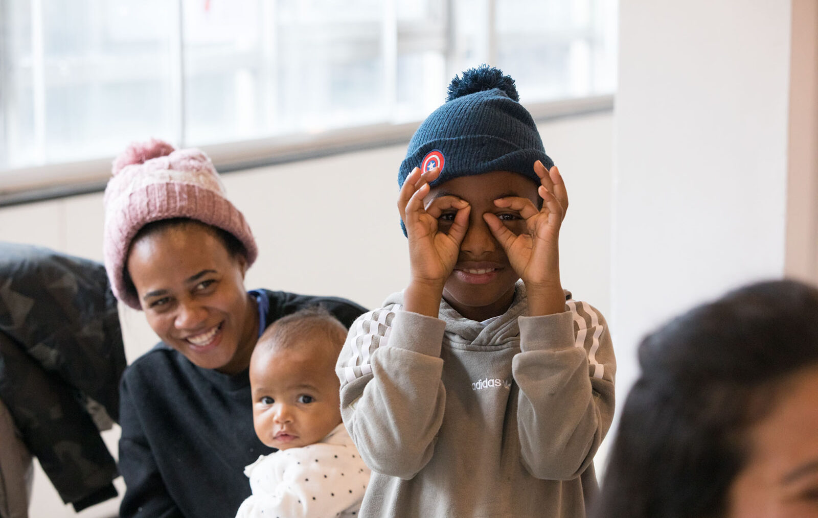 A young black boy with a blue bobble hat and grey jumper stands smiling, holding his hands like glasses over his eyes.