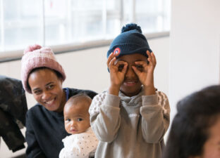 A young black boy with a blue bobble hat and grey jumper stands smiling, holding his hands like glasses over his eyes.