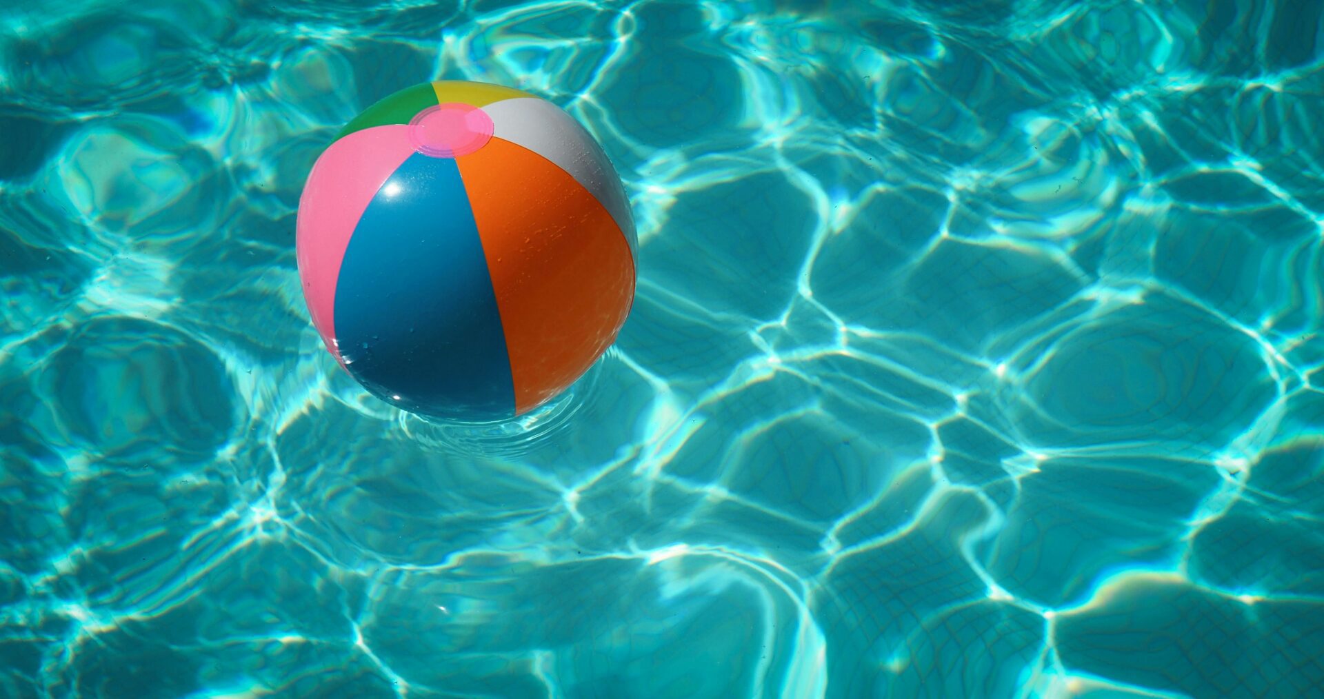 A colourful striped beach ball floats on top of bright blue swimming pool water.