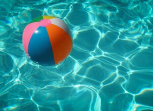 A colourful striped beach ball floats on top of bright blue swimming pool water.