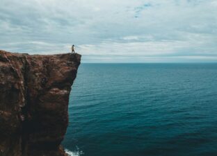 A persons stands atop a cliff, jutting out over the blue ocean below