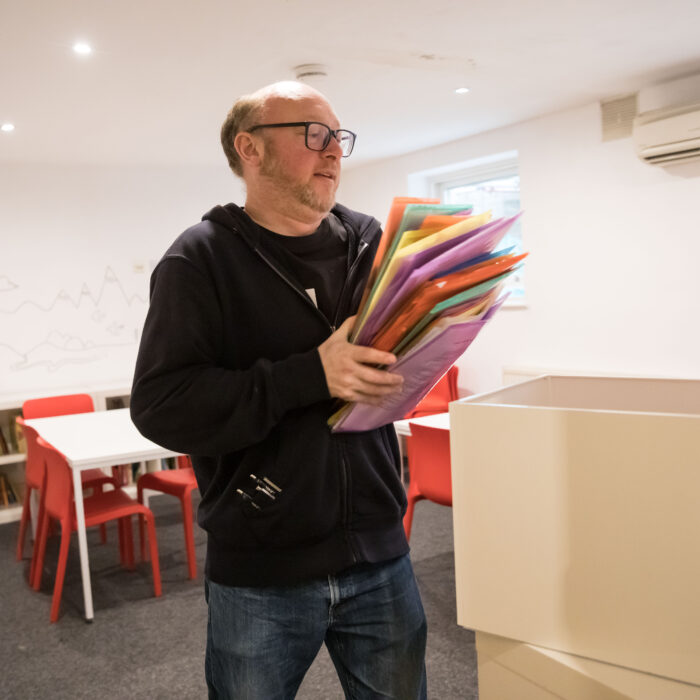 In this photograph one of writing mentors gathers a colourful stack of folders. He is white, with very short hair and is wearing glasses. His clothes are dark and he is looking at the folders. 