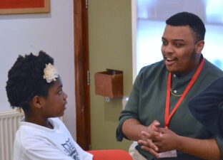 Ashanté - a young black man in a green button up shirt - sits and chats to a young writer with a flower in her hair. They are in the Ministry of Stories workshop space.