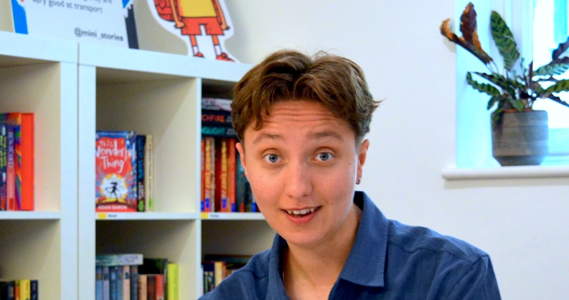 Beattie is a young, white person with short brown hair. They're sitting behind a desk wearing a blue short sleeved shirt and smiling at the camera. Their hands are folded on the desk in front of them.