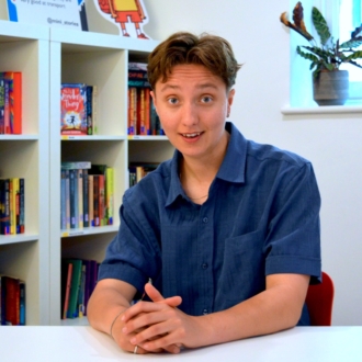 Beattie is a young, white person with short brown hair. They're sitting behind a desk wearing a blue short sleeved shirt and smiling at the camera. Their hands are folded on the desk in front of them.