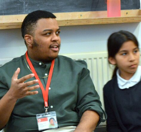 Ashanté, a young black man, with white torusers and a green shirt sits on the floor of the Ministry smiling and chatting with young children about their writing.