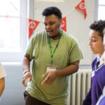 Ashanté - a young man in a green t-shirt - gesticulates as he leads a workshop for secondary school children. There is Ministry of Stories bunting behind him in the hall of a school. You can see two pupils, both in purple school uniforms.
