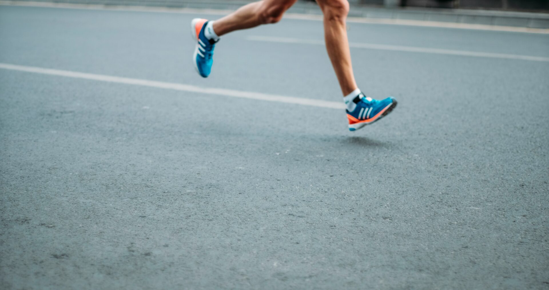 a runner runs down a grey concrete road, we see there legs and feet in blue and orange trainers.