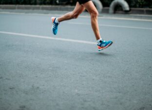 a runner runs down a grey concrete road, we see there legs and feet in blue and orange trainers.