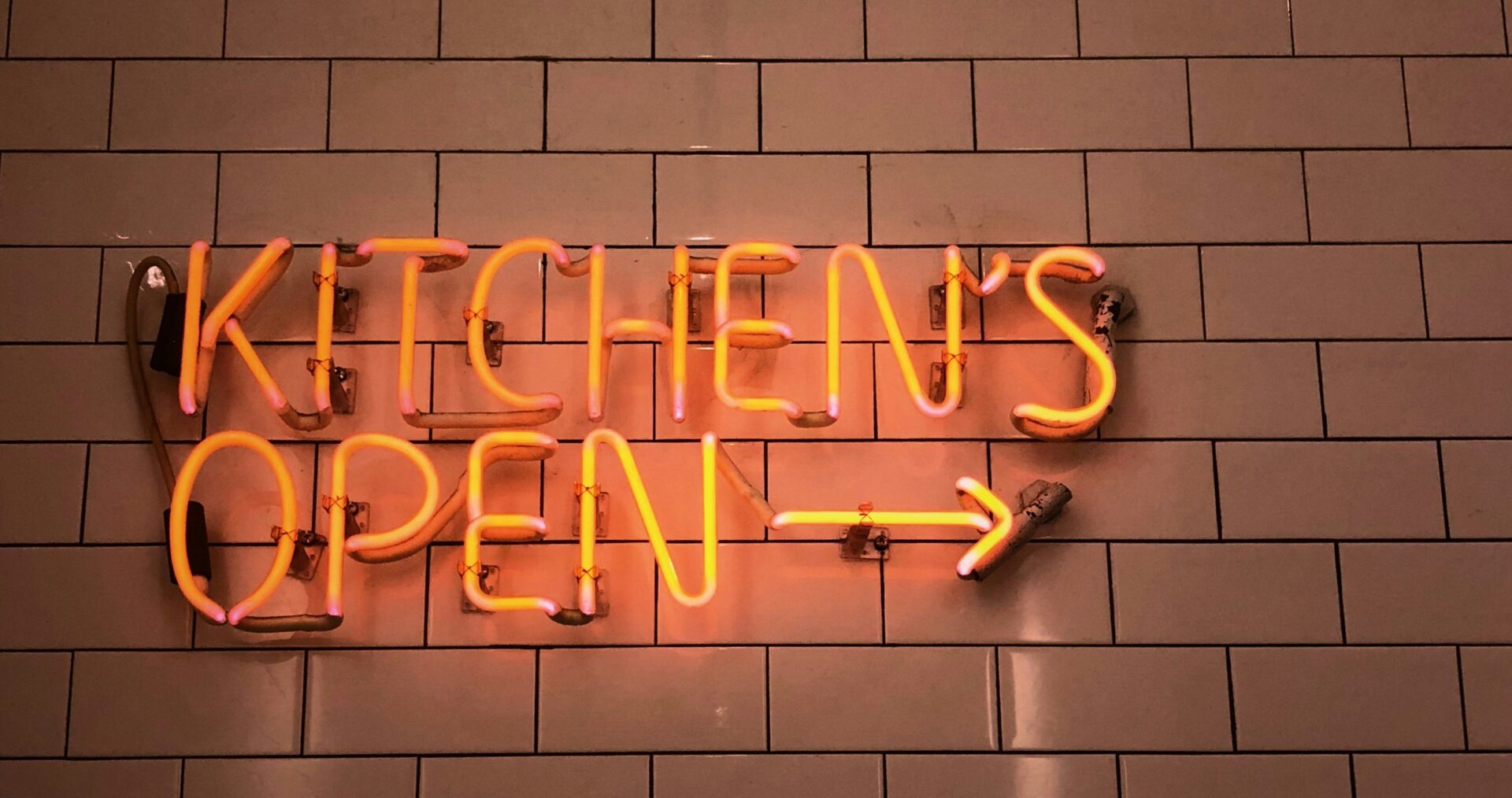 A bright red neon sign reads 'kitchen is open' with a glowing arrow pointing along a red brick wall.