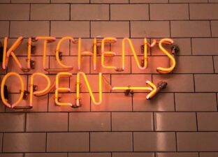 A bright red neon sign reads 'kitchen is open' with a glowing arrow pointing along a red brick wall.
