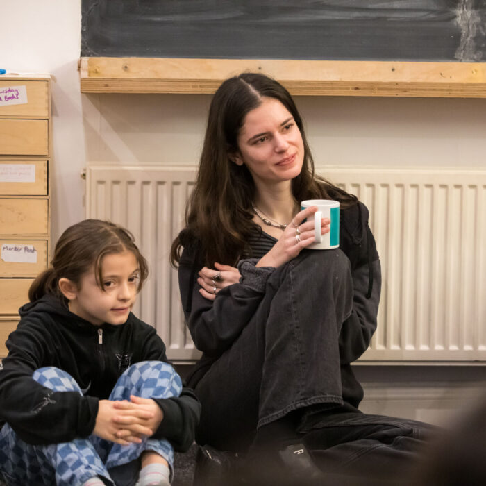 This photograph shows a volunteer sitting on the carpet with young writers at the Ministry of Stories writing centre. She is white, has long brown hair, and is wearing dark colours. She is holding a cup of tea which she balances on her knee. to her left, a young writer is sitting on the carpet too, wearing chequered jeans and a dark top. They are both listening attentively to the writing facilitator who is out of frame. 