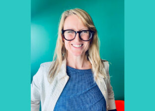 Rebecca - a white woman, with long blonde hair and bold, black glasses - sits grinning broadly at the camera. She wears a blue top with cream jacket and sits in front of a teal green wall.