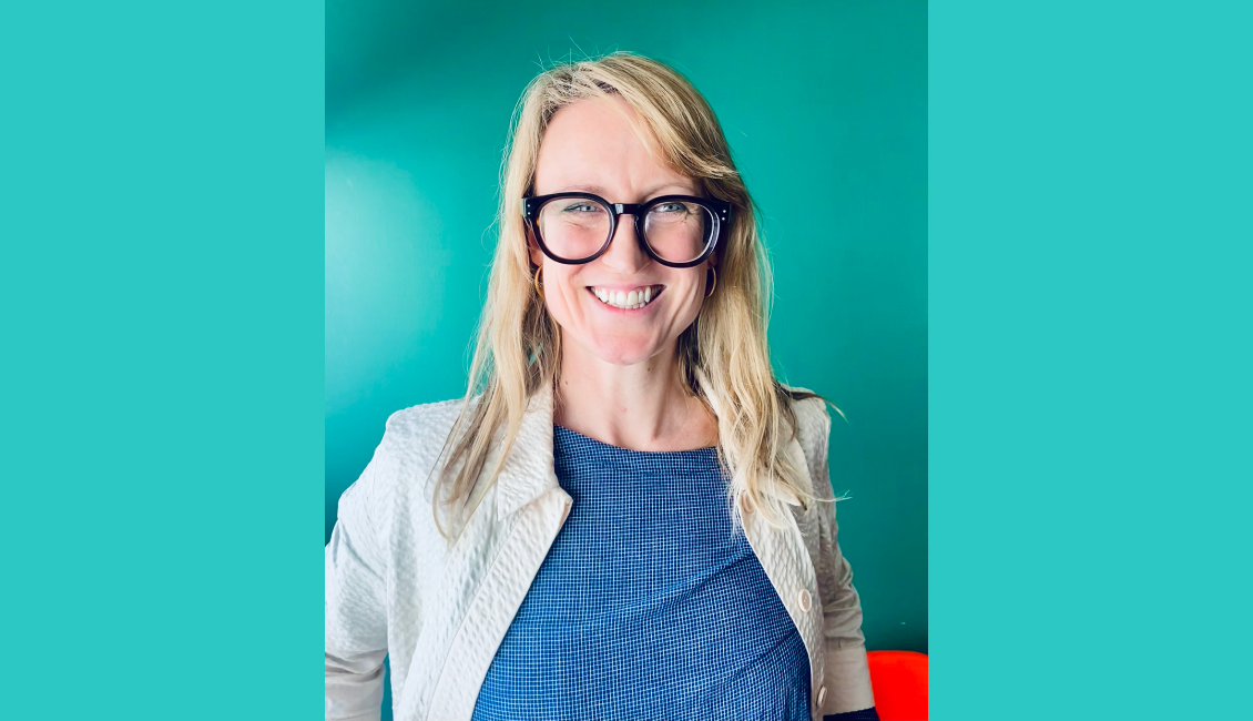 Rebecca - a white woman, with long blonde hair and bold, black glasses - sits grinning broadly at the camera. She wears a blue top with cream jacket and sits in front of a teal green wall.