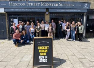 A large group of teachers stand cheering in front of the store front Hoxton Street Monster Supplies.