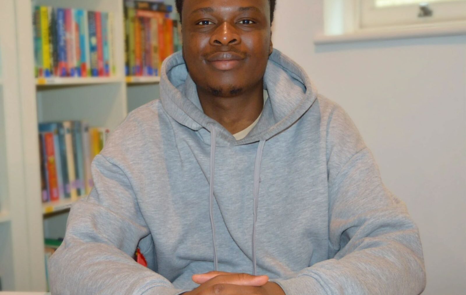 Joshua is a young black man with short hair. He is sat down and facing the camera, with his hands clasped in front of him on a white table. Behind him is a book shelf with colourful books on it. He is wearing a grey hoodie and smiling.