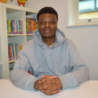 Joshua is a young black man with short hair. He is sat down and facing the camera, with his hands clasped in front of him on a white table. Behind him is a book shelf with colourful books on it. He is wearing a grey hoodie and smiling.