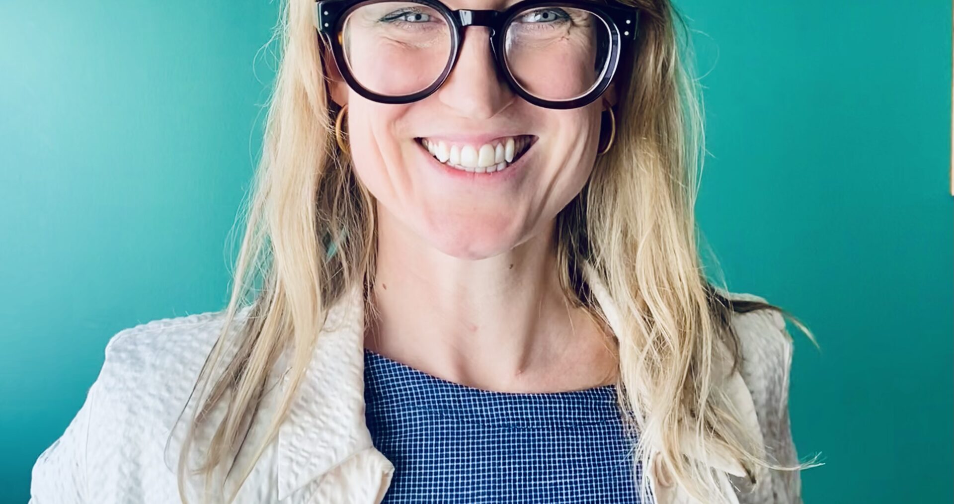 Rebecca - a white woman, with long blonde hair and bold, black glasses - sits grinning broadly at the camera. She wears a blue top with cream jacket and sits in front of a teal green wall.