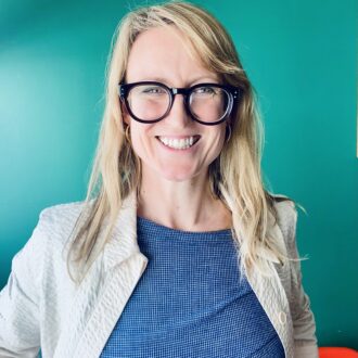 Rebecca - a white woman, with long blonde hair and bold, black glasses - sits grinning broadly at the camera. She wears a blue top with cream jacket and sits in front of a teal green wall.