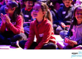 5 or so primary school children in school uniforms are sitting cross-legged on the floor of a dramatically lit auditorium. They are smiling and clutching small goody bags. The Amazon Literary Partnership Logo appears at the bottom right on a white banner that runs underneath the image.