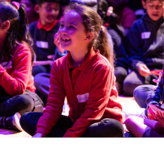 5 or so primary school children in school uniforms are sitting cross-legged on the floor of a dramatically lit auditorium. They are smiling and clutching small goody bags. The Amazon Literary Partnership Logo appears at the bottom right on a white banner that runs underneath the image.